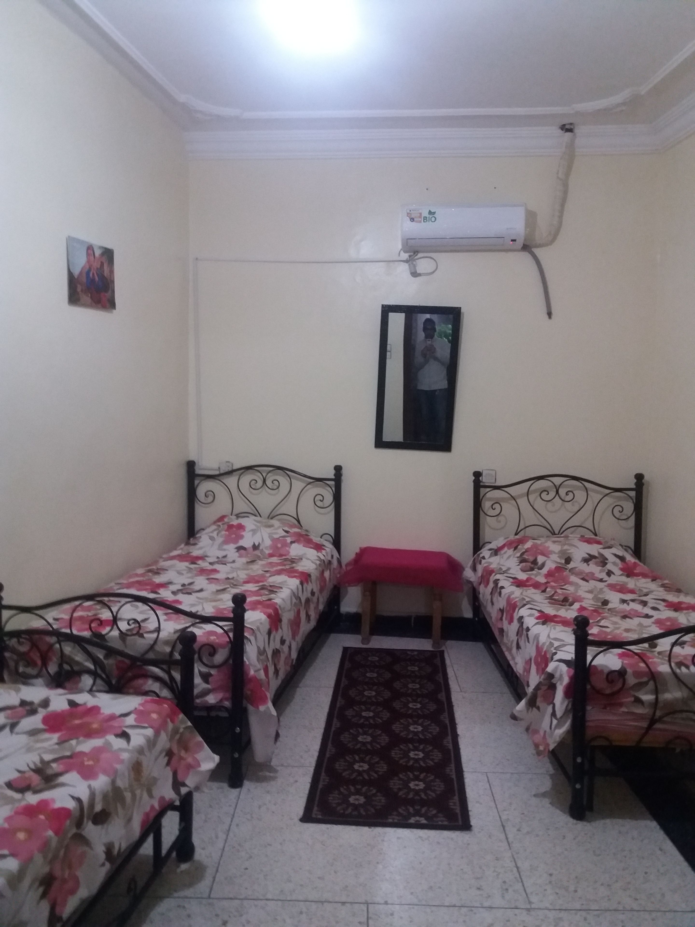 A room with two single beds, a small bench, a mirror, and a wall-mounted air conditioner.