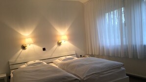 A double bed with white linens in a room with two wall-mounted lamps and a window with sheer curtains. - Hotel Wintersmühle (Bielefeld)