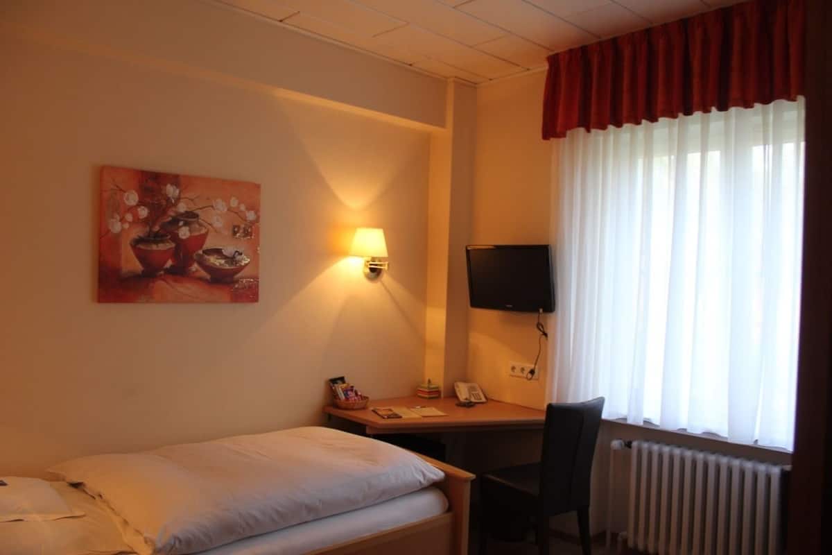 Habitación de hotel con cama, escritorio con silla, televisor, lámpara y ventana con cortinas.