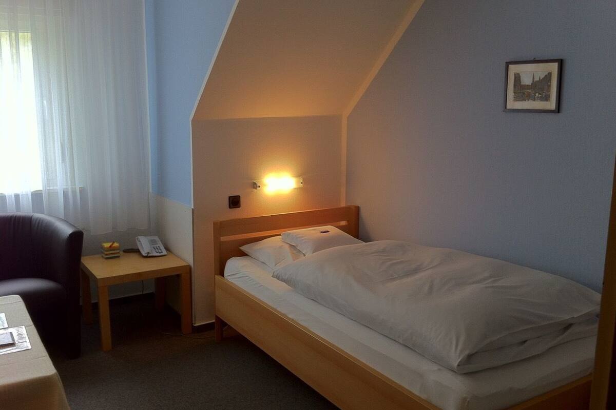 Una habitación pequeña con cama individual, techo inclinado, mesita de noche con lámpara, una silla y un escritorio con teléfono.