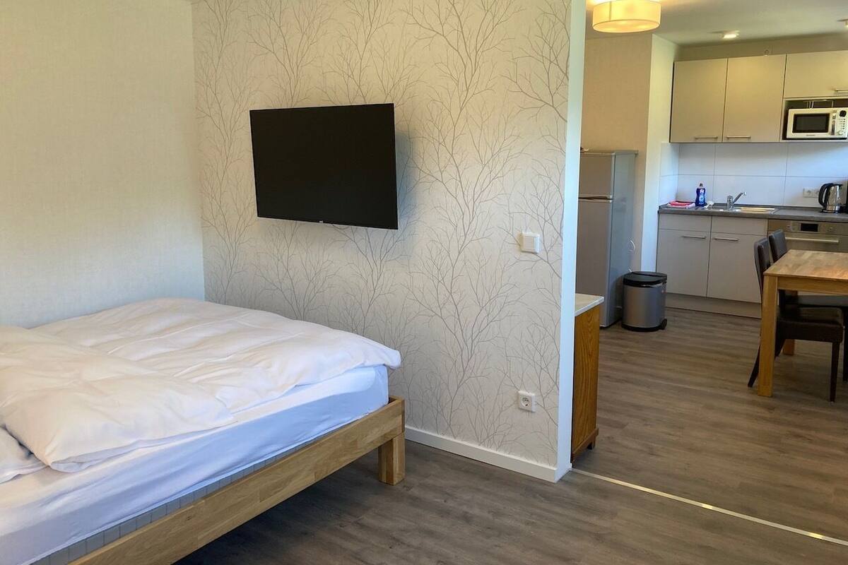 Una habitación compacta con una cama, un televisor montado en la pared, una kitchenette con microondas y una zona de comedor con mesa y sillas.