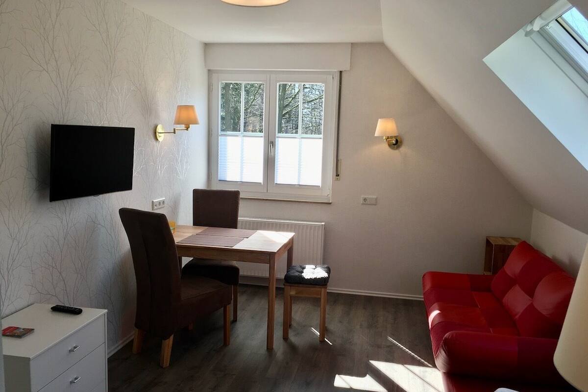 Una habitación con una mesa de comedor, sillas, un sofá rojo, un televisor montado en la pared y un ventanal con vistas a árboles.