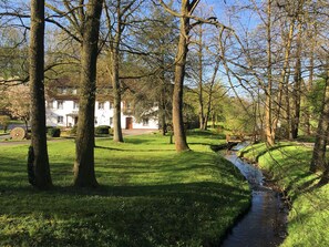 A serene park with a small stream, lush green grass, and a white building with a red door in the background. - Hotel Wintersmühle (Bielefeld)