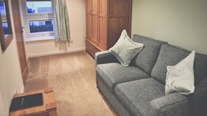 A room with a grey sofa, two white pillows, a wooden wardrobe, and a window with curtains. - Count House Cottage B&B - Adults Only (St Ives)