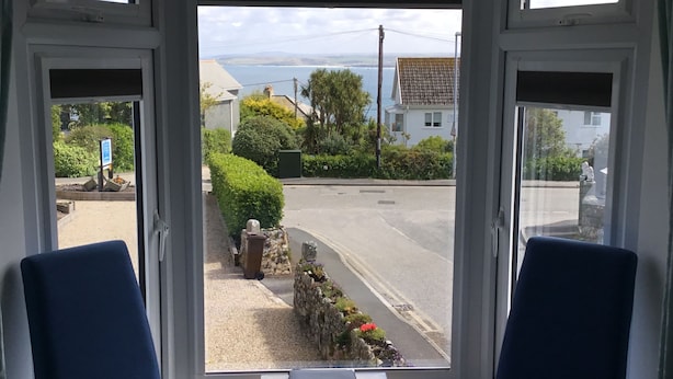 A room with two blue chairs facing a window overlooking a street with houses, a road, and a body of water in the distance. - Count House Cottage B&B - Adults Only (St Ives)