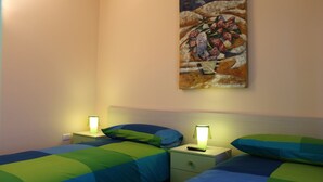 Quarto de hotel com duas camas, cada uma com manta verde e azul, mesa de cabeceira com abajur e um quadro na parede.
