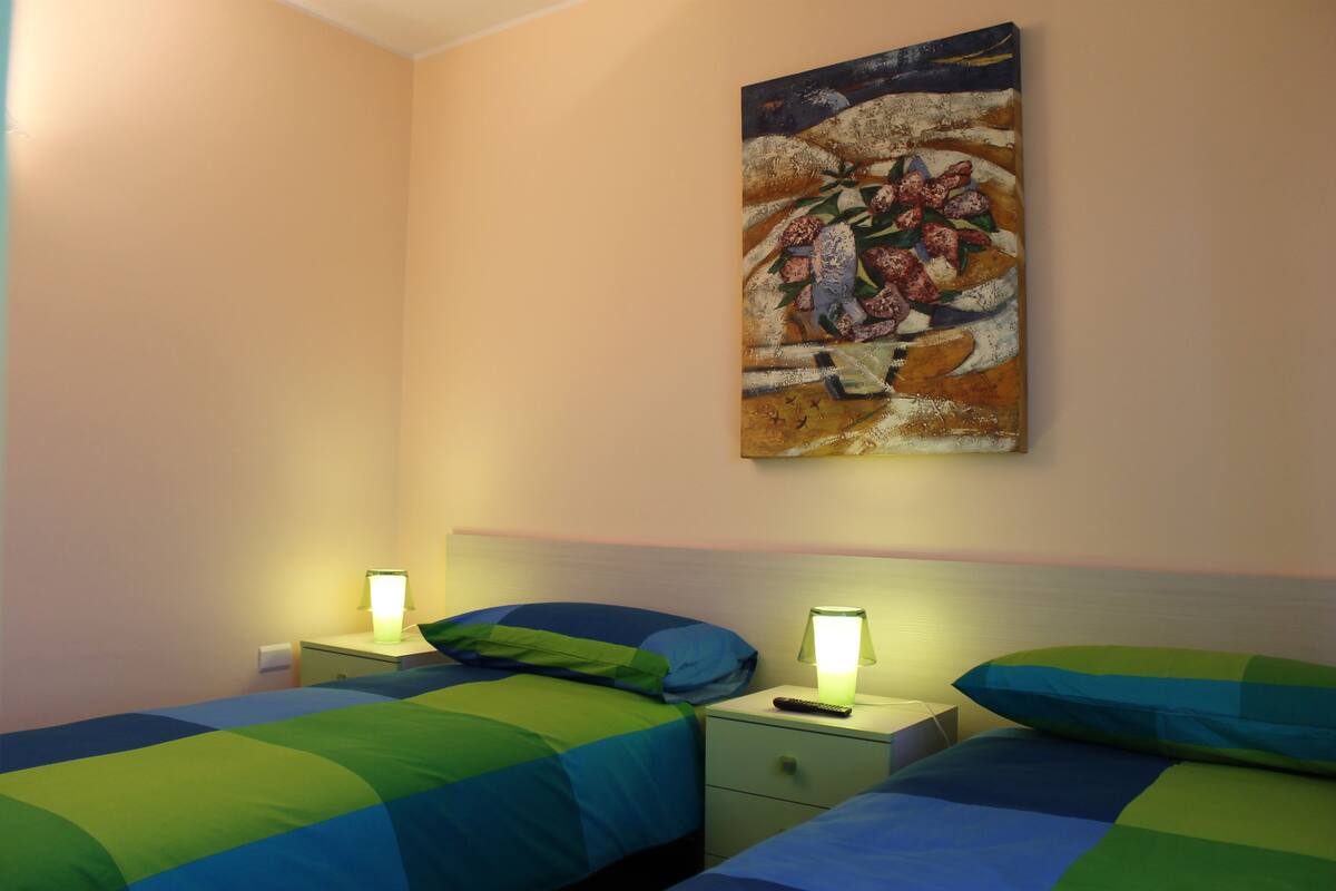 Una habitación de hotel con dos camas, cada una con colcha verde y azul, una mesita de noche con lámpara y un cuadro en la pared.