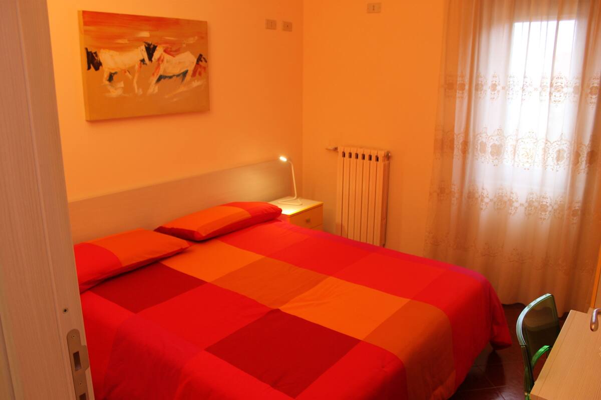 Una habitación con cama, mesita de noche, silla, un cuadro en la pared y ventana con cortinas.