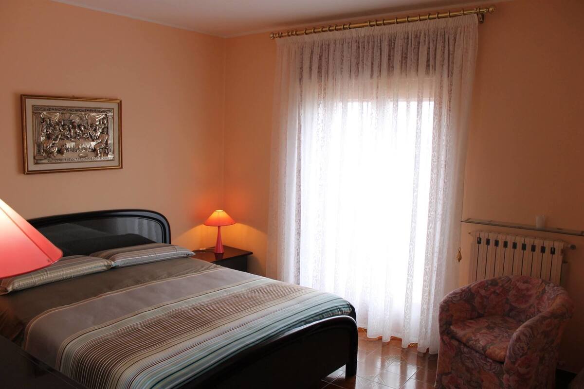 Un dormitorio con cama, mesita de noche con lámpara, una silla, radiador y ventana con cortinas traslúcidas.