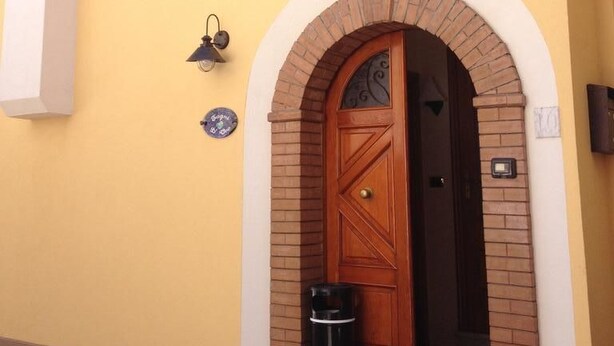 Um edifício amarelo com porta de madeira sob uma entrada em arco, uma pequena lixeira e um aplique na parede.