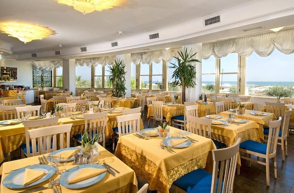 A restaurant with multiple tables set for dining, large windows offering a view of the sea, and a bar area visible in the background.