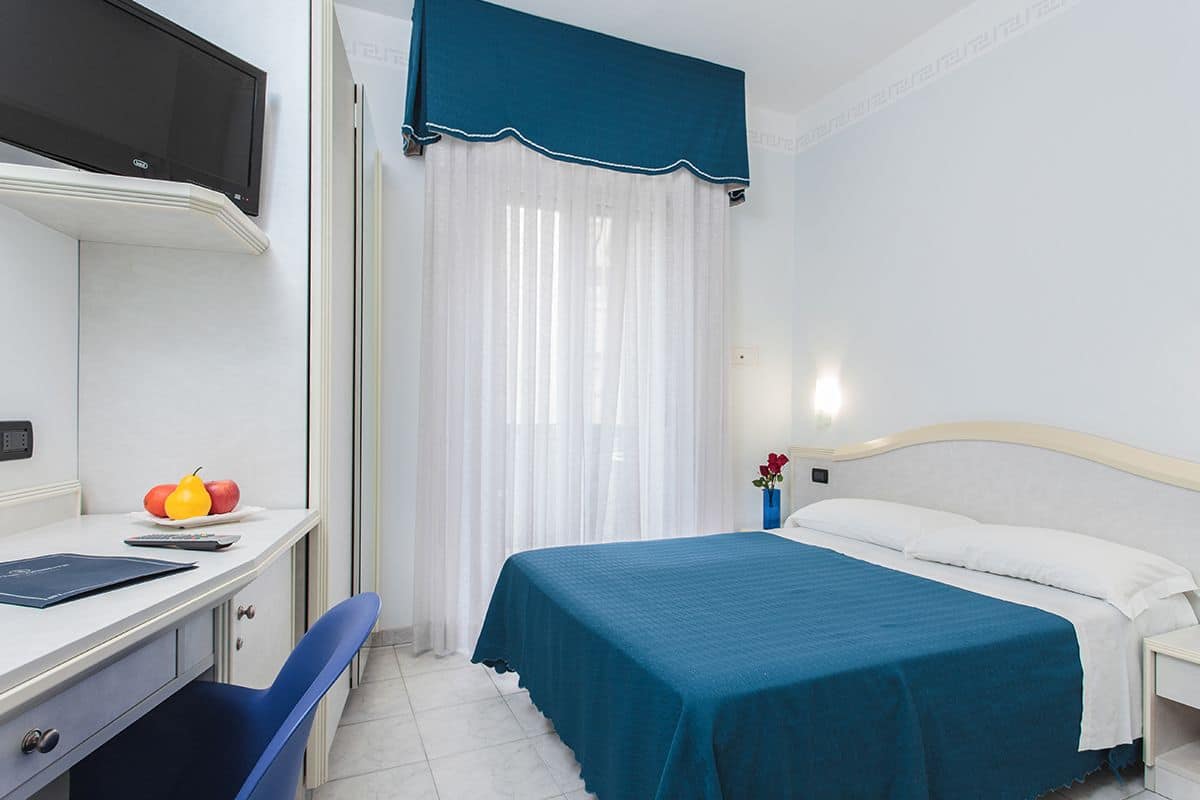 A hotel room with a bed, a desk with a chair, a TV, and a window with curtains.