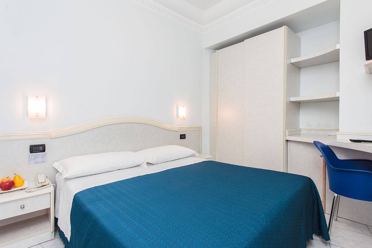 A hotel room with a bed, a desk, and a chair. There is a nightstand with a phone and a fruit plate.