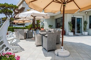 An outdoor patio area with multiple tables and chairs, surrounded by umbrellas and potted plants. - Hotel Oriente (Cervia)