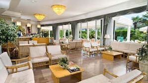 A spacious lounge area with wicker furniture, a central coffee table, and large windows offering a view of the outdoors. - Hotel Oriente (Cervia)
