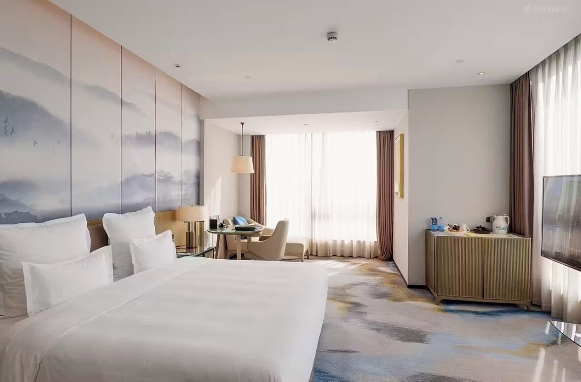 A hotel room with a large bed, a desk with a chair, a small table, a TV, and a window with curtains.