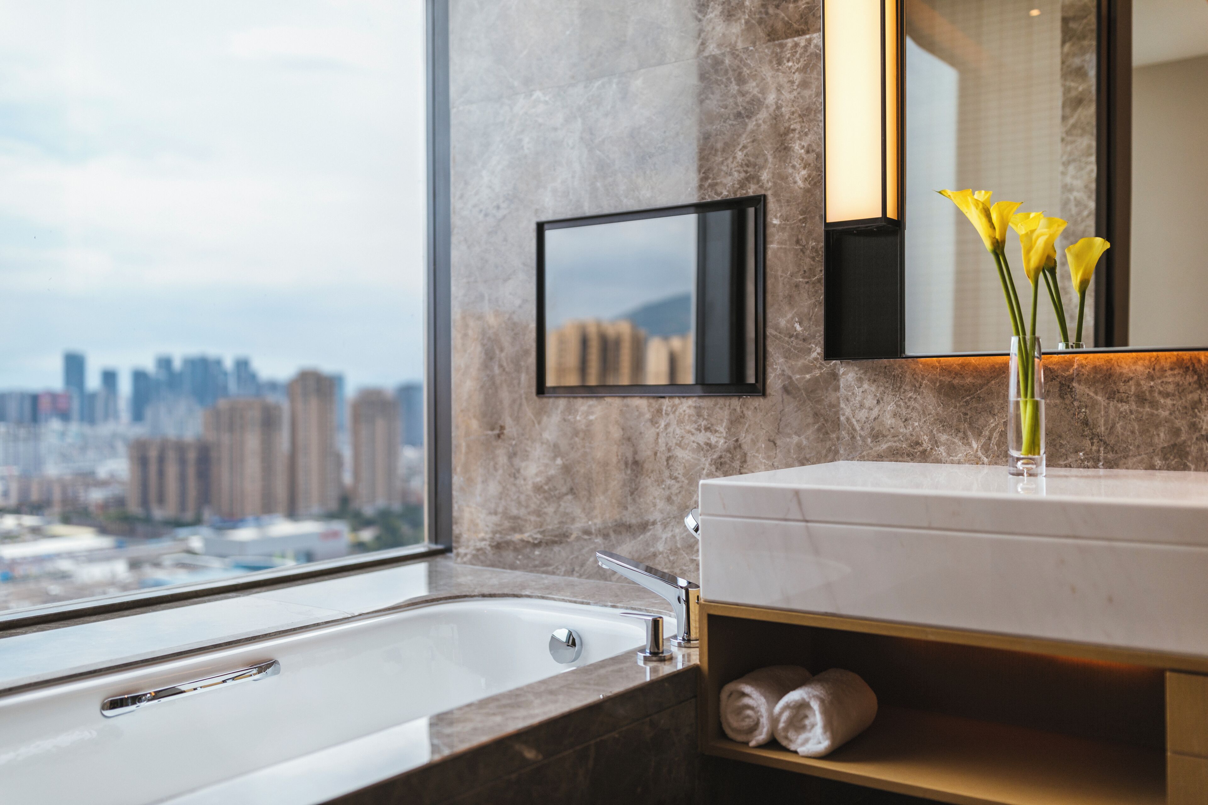 A modern bathroom with a large window, a bathtub, a mirror, and a vase with flowers.