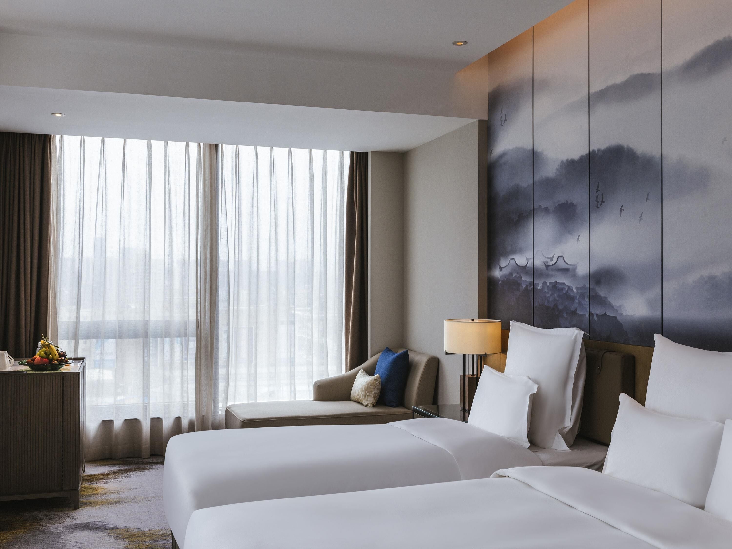 Habitación de hotel con dos camas, un escritorio, una lámpara y un amplio ventanal con cortinas.