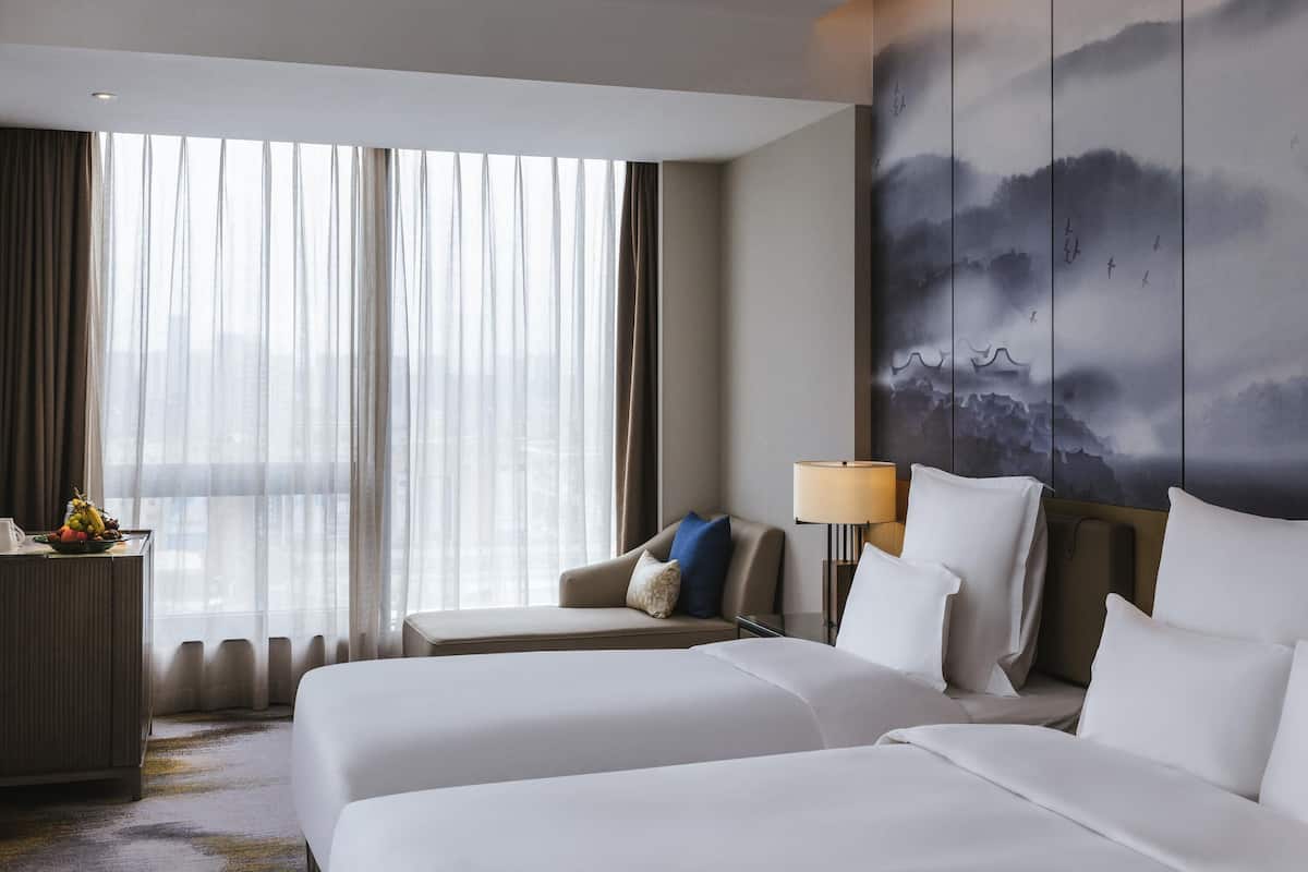 Ein Hotelzimmer mit zwei Betten, einem Schreibtisch, einer Lampe und einem großen Fenster mit Vorhängen.