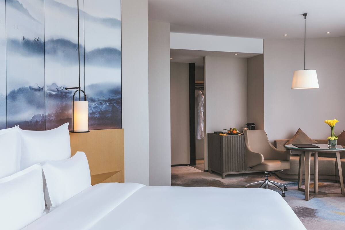 Ein modernes Hotelzimmer mit einem großen Bett, einem Schreibtisch, einem Sessel und einem Gemälde an der Wand.