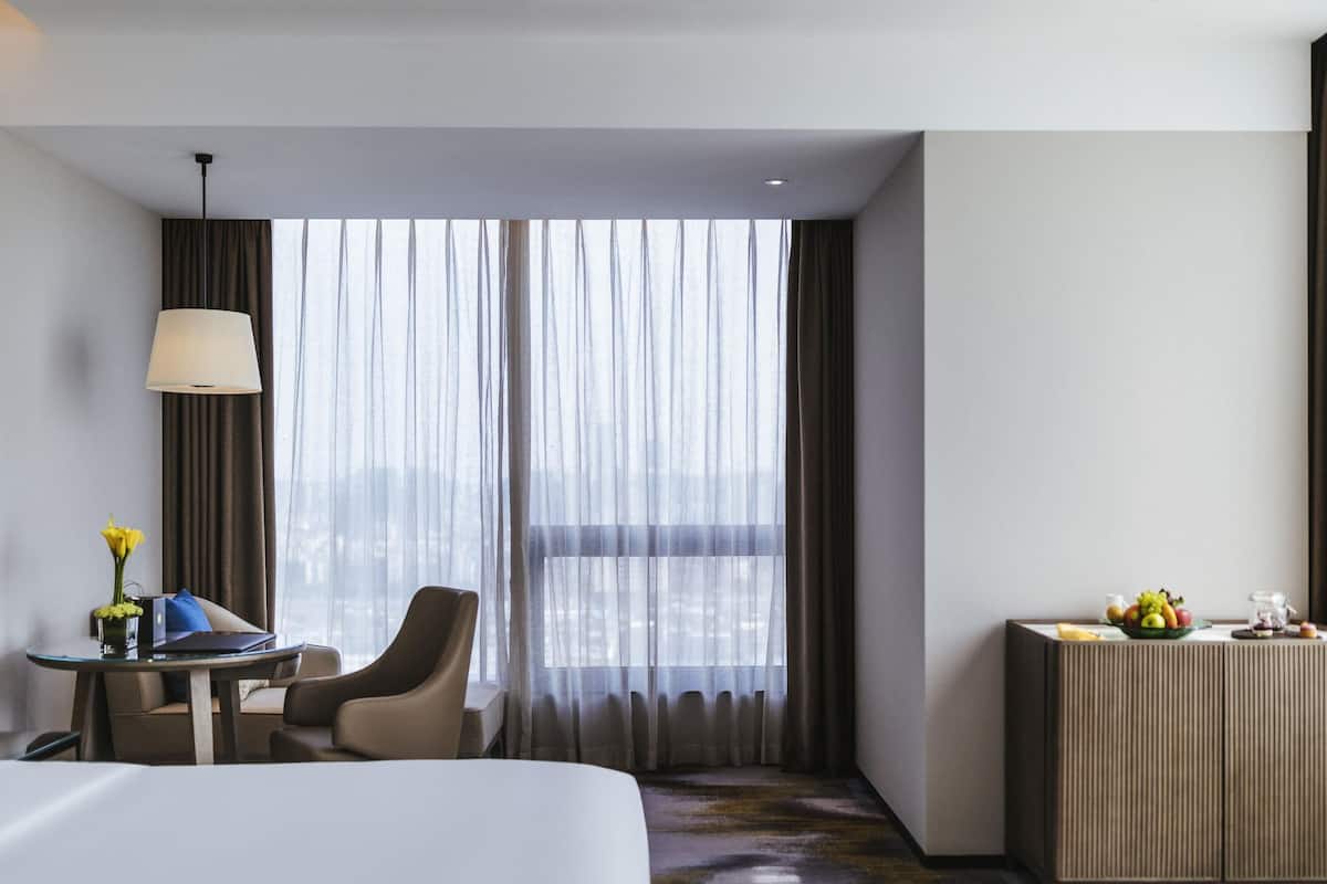 Ein Hotelzimmer mit einem großen Bett, einem Schreibtisch mit Lampe, einem Sessel, einem kleinen Tisch mit Blumen und Blick auf die Stadt durch leichte Vorhänge.