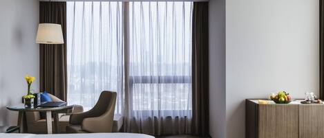 Une chambre d’hôtel avec un grand lit, un bureau avec une lampe, une chaise, une petite table avec un vase de fleurs et une vue sur la ville à travers de fines rideaux.