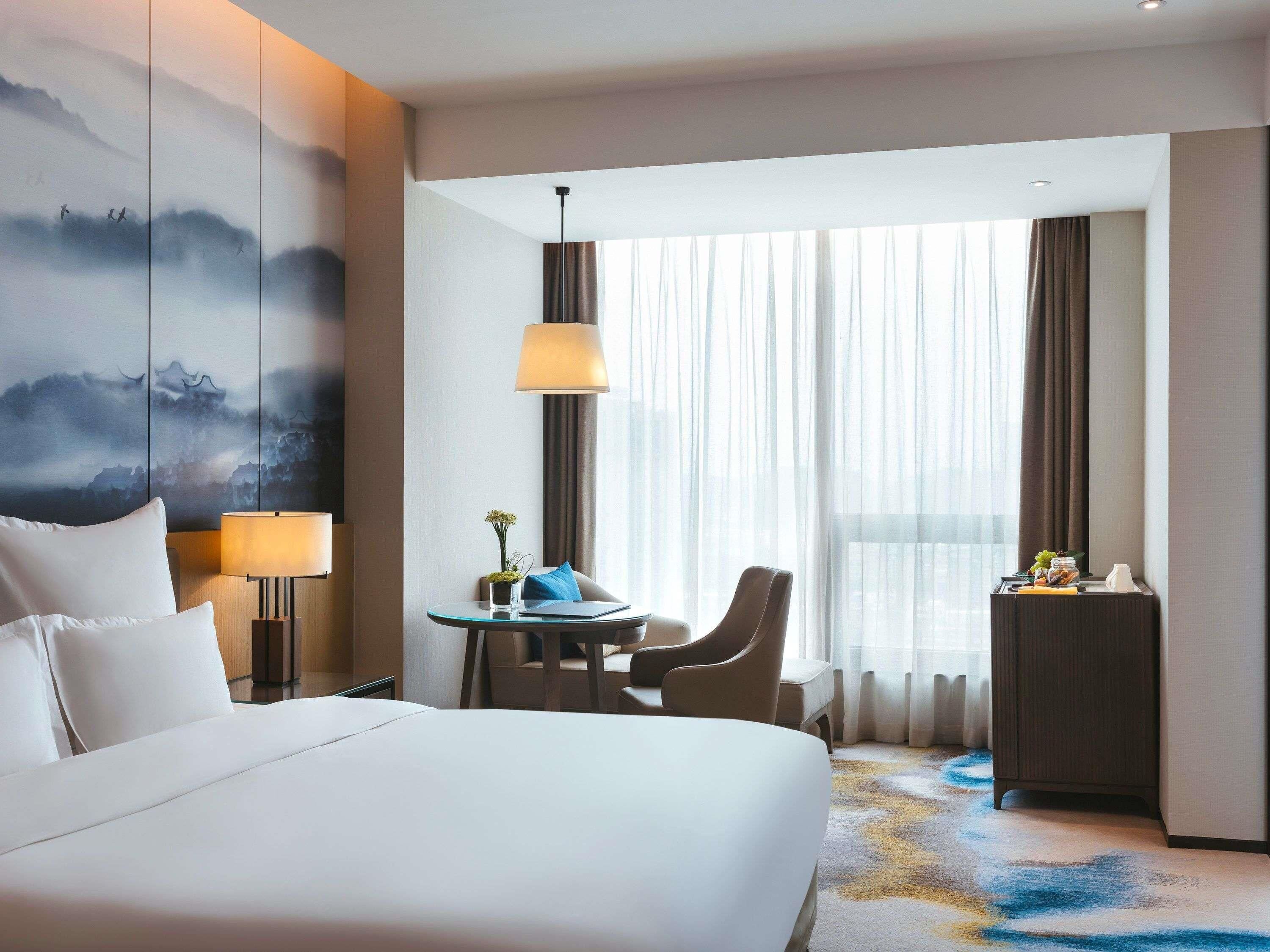 Habitación de hotel con una cama grande, un escritorio con silla, una lámpara y vistas a la ciudad a través de cortinas translúcidas.