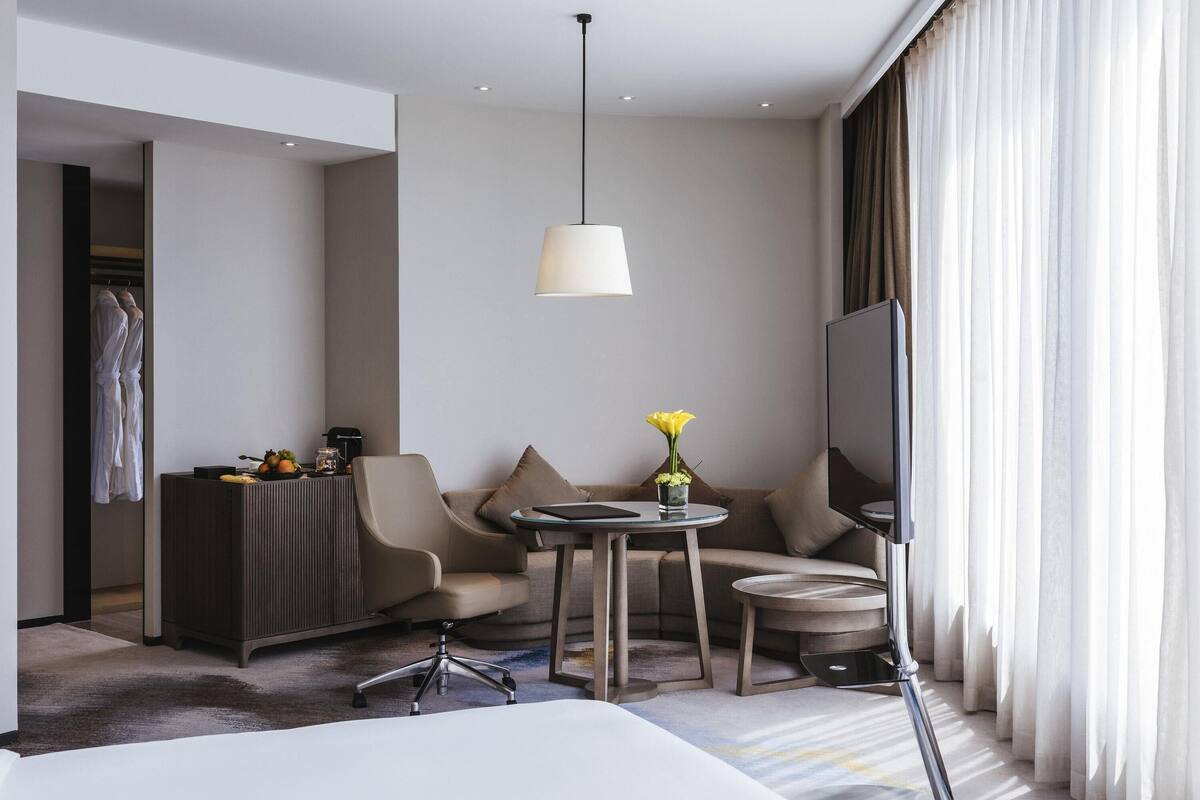 Ein modernes Hotelzimmer mit Bett, Schreibtisch, Stuhl, einem kleinen Tisch mit einer Vase, einem Fernseher und einem Kleiderschrank.