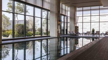A modern indoor swimming pool with large windows, lounge chairs, and a clear view of the surrounding greenery.