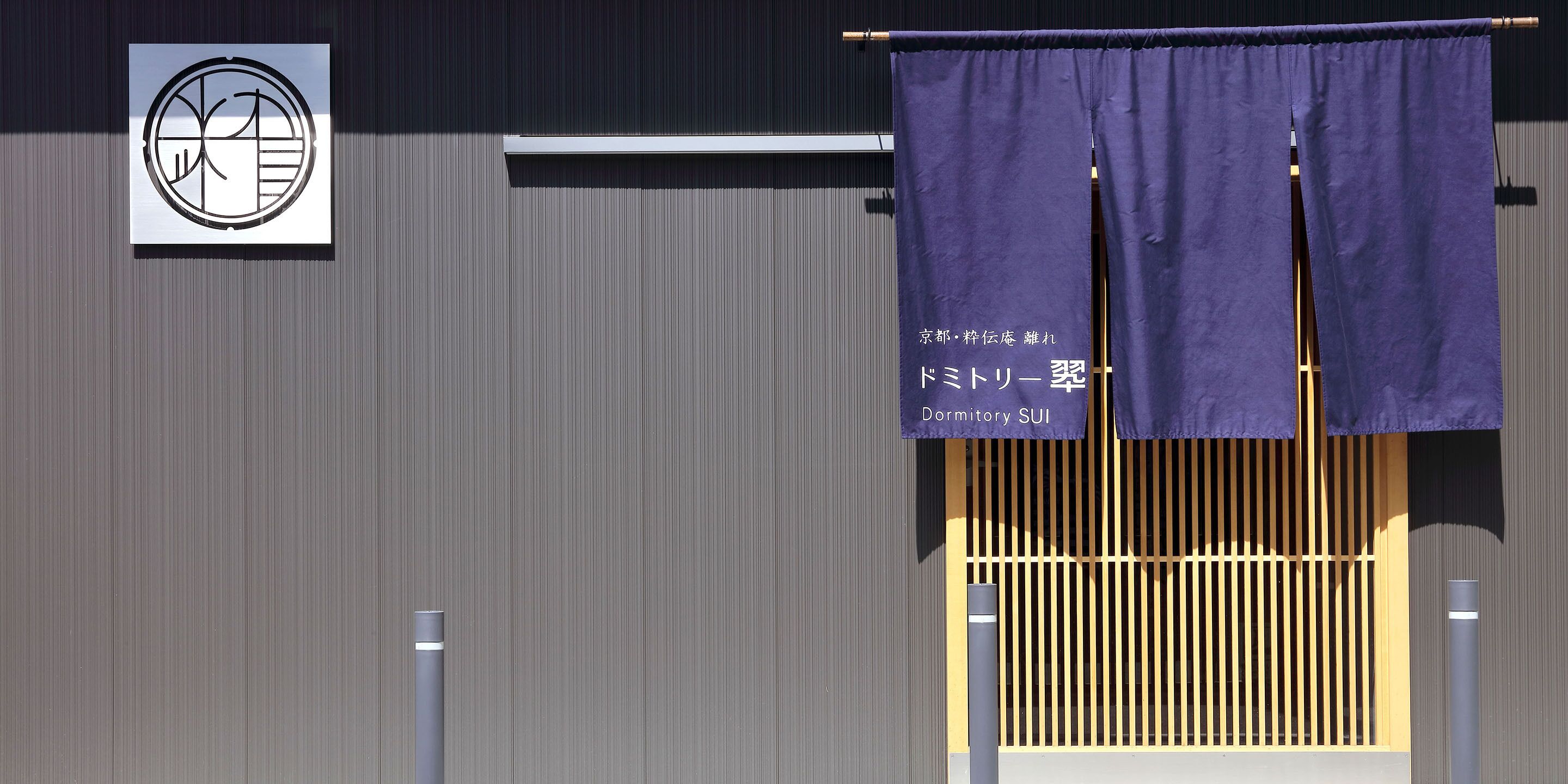 Fachada de edificio con un estampado a rayas verticales, un letrero con caracteres japoneses y un banderín morado con texto.