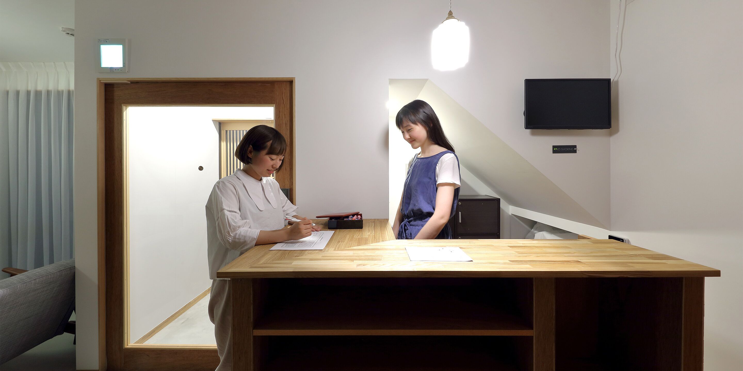 A room with a wooden desk, a chair, a bed, a TV mounted on the wall, and a window with blinds.