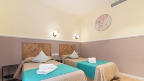 Habitación de hotel con dos camas, cada una con una colcha color turquesa y una toalla blanca sobre la cama. Hay luces fijadas a la pared y un cuadro redondo en ella.