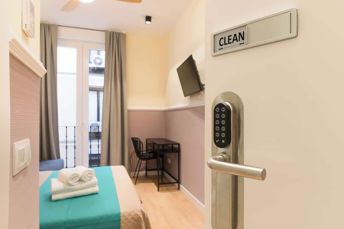 Quarto de hotel com cama, escrivaninha, cadeira, televisão presa à parede e porta com a placa 'CLEAN'.