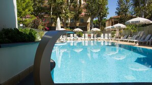 Una piscina con sillones de descanso y sombrillas, rodeada de vegetación y edificios.