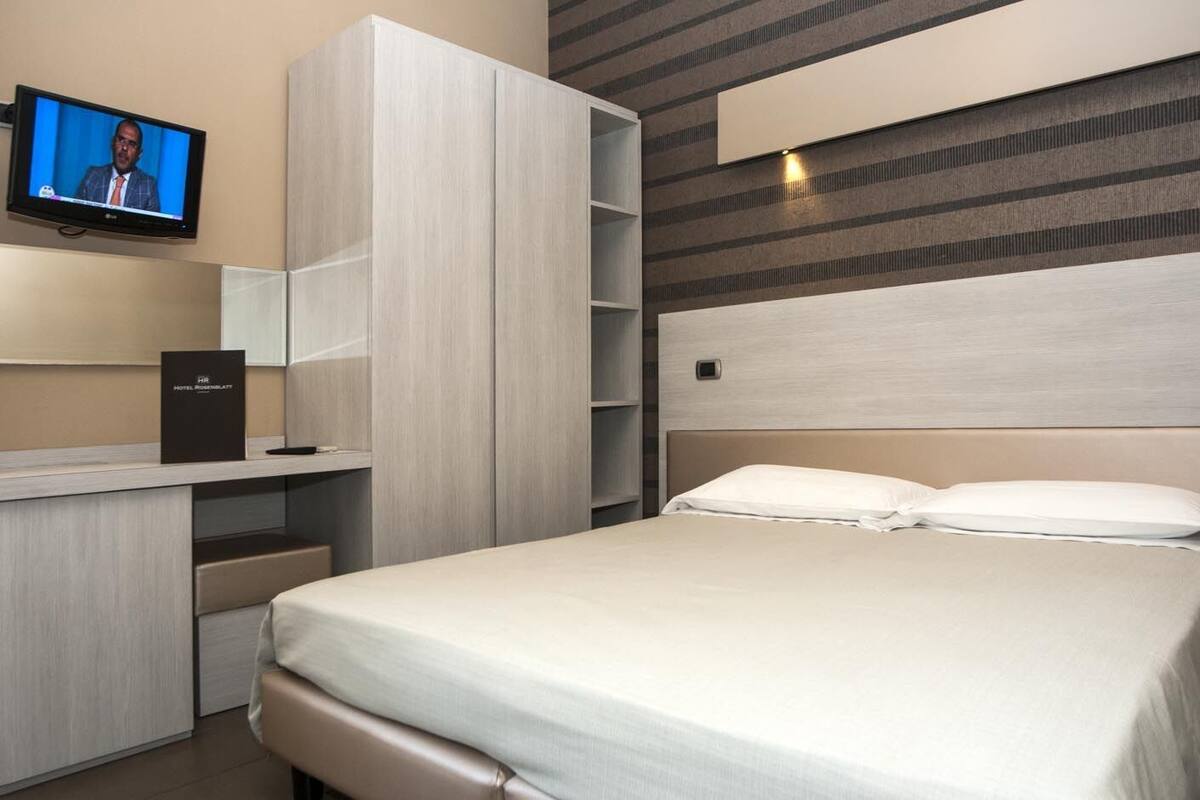 A modern hotel room with a bed, a TV mounted on the wall, a desk with a computer, and a bookshelf.