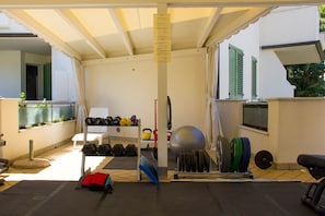 A covered outdoor gym area with exercise equipment including dumbbells, kettlebells, a medicine ball, and a bench press. - Hotel Rosenblatt (Cervia)