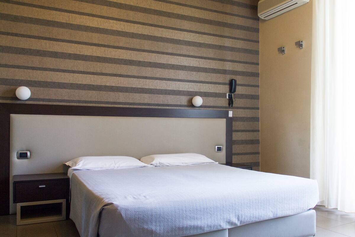 A hotel room with a bed, bedside tables, a wall-mounted air conditioner, and a window with curtains.