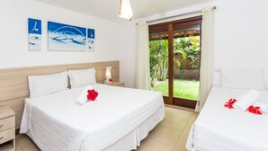 A hotel room with two beds, a wooden headboard, a nightstand with a lamp, and a view of greenery through a sliding glass door. - Toko Village (Porto Seguro)
