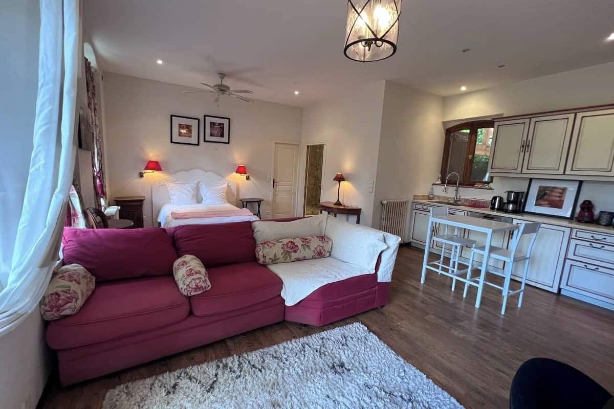A bedroom with a bed, a red sofa, a white armchair, a small table, and a kitchenette.