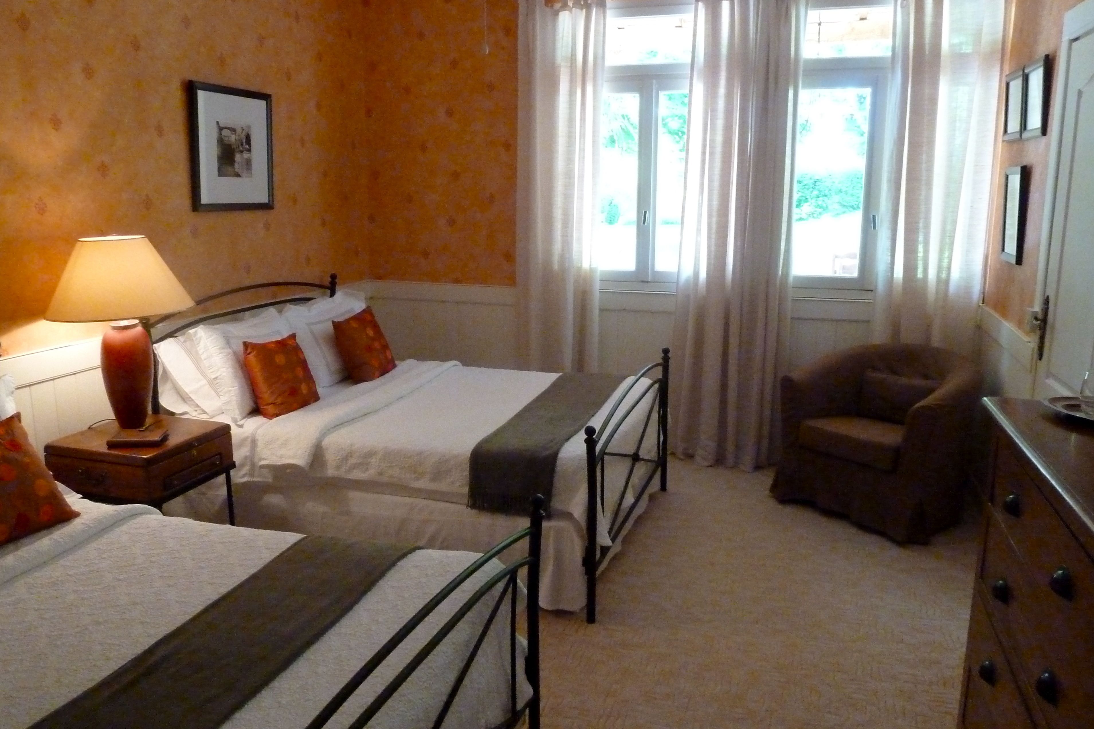 Habitación de hotel con dos camas, una silla, un aparador, una lámpara y una ventana con cortinas.