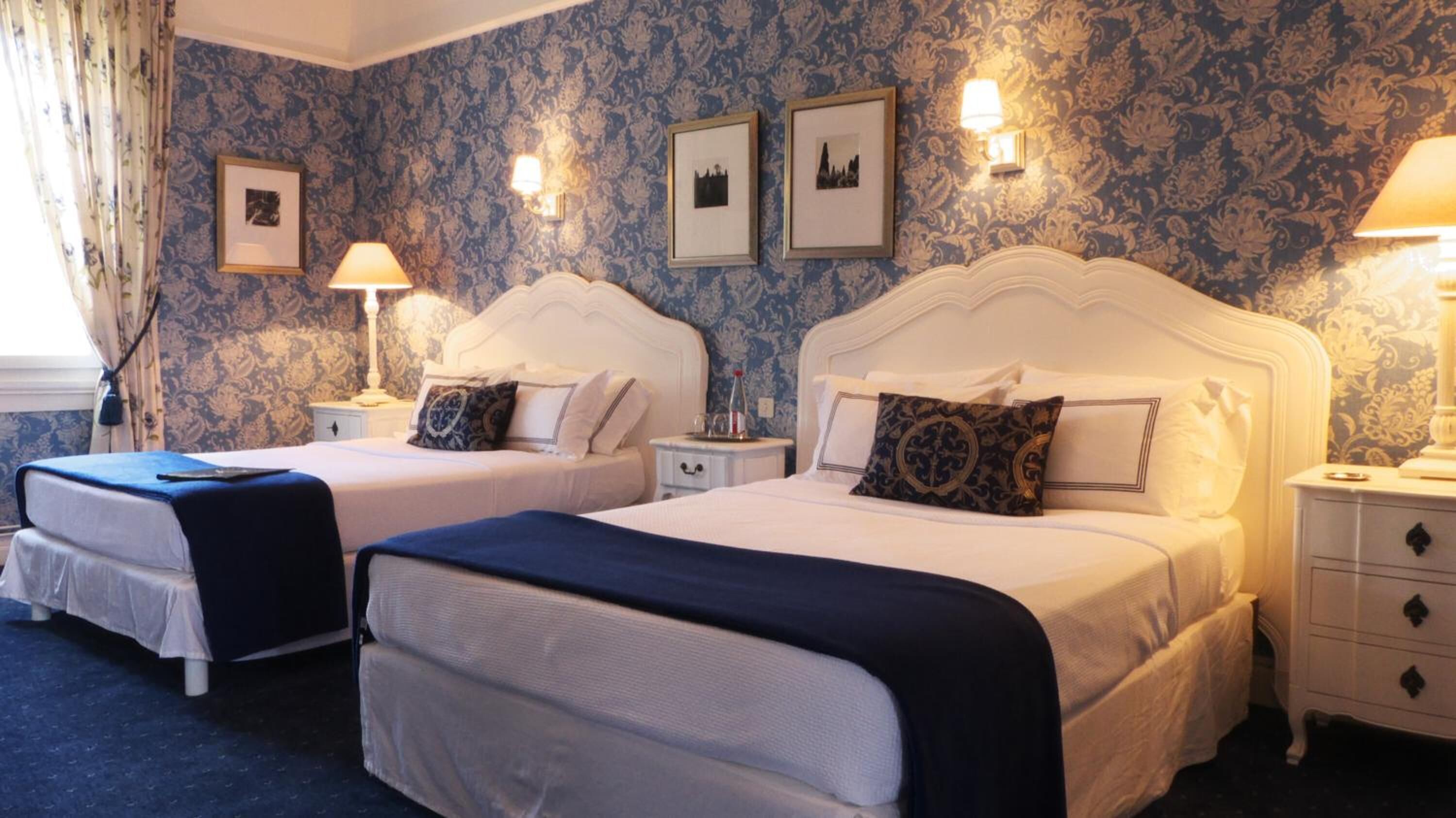 A hotel room with two beds, a nightstand, a lamp, and a wall with framed pictures.