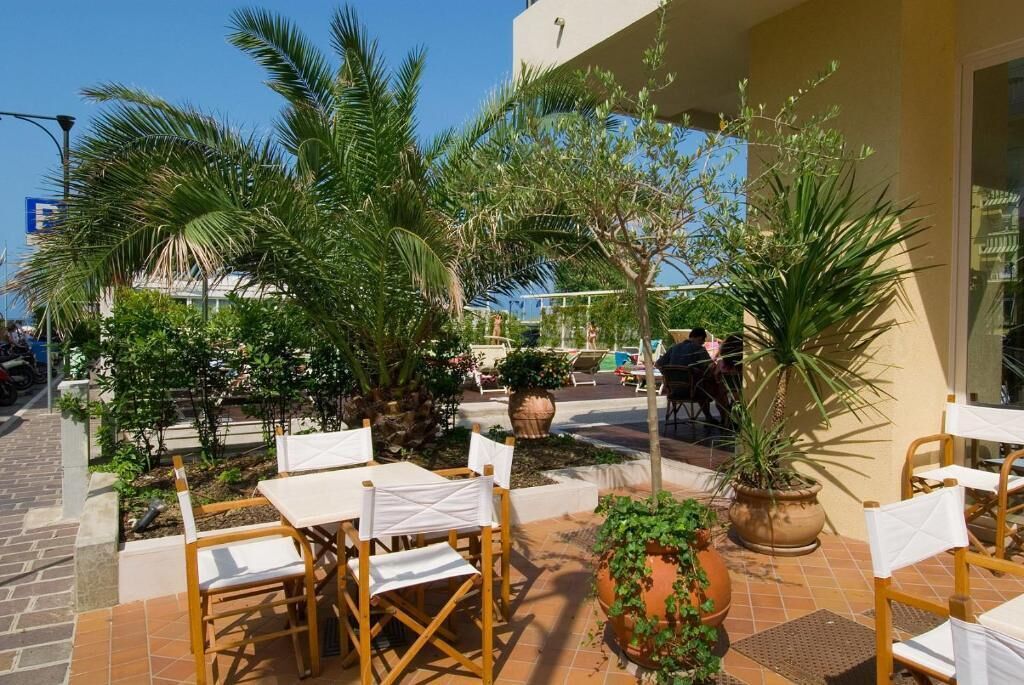 An outdoor patio area with tables and chairs, surrounded by potted plants and palm trees.