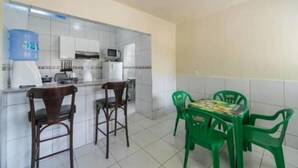 Una pequeña kitchenette con microondas, cafetera y dispensador de agua. Hay dos sillas de madera y una mesa con sillas de plástico verde.