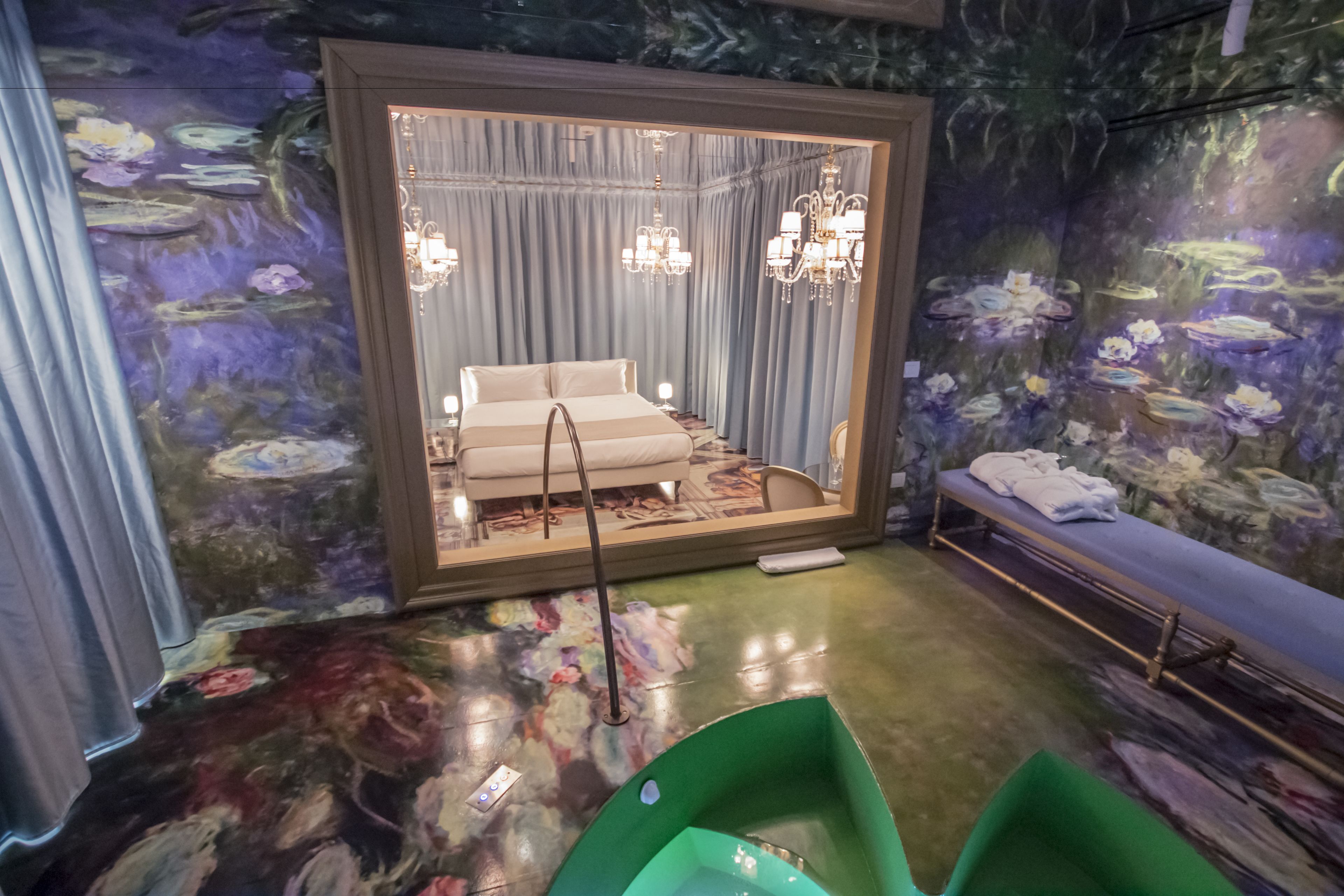 A room with a mirrored wall, a bed, a bench, and a green chair.