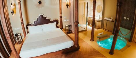 A junior suite with a bed, wooden pillars, a small table, a chair, a hanging light fixture, and a pool area.