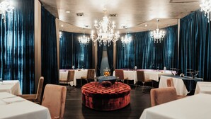 Un restaurant avec des tables rondes, des rideaux en velours, des lustres et un ottoman central.