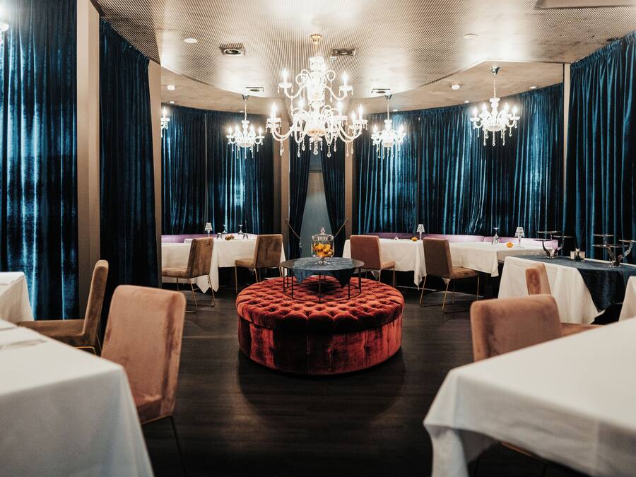 Een restaurant met ronde tafels, fluwelen gordijnen, kroonluchters en een centraal geplaatste poef.