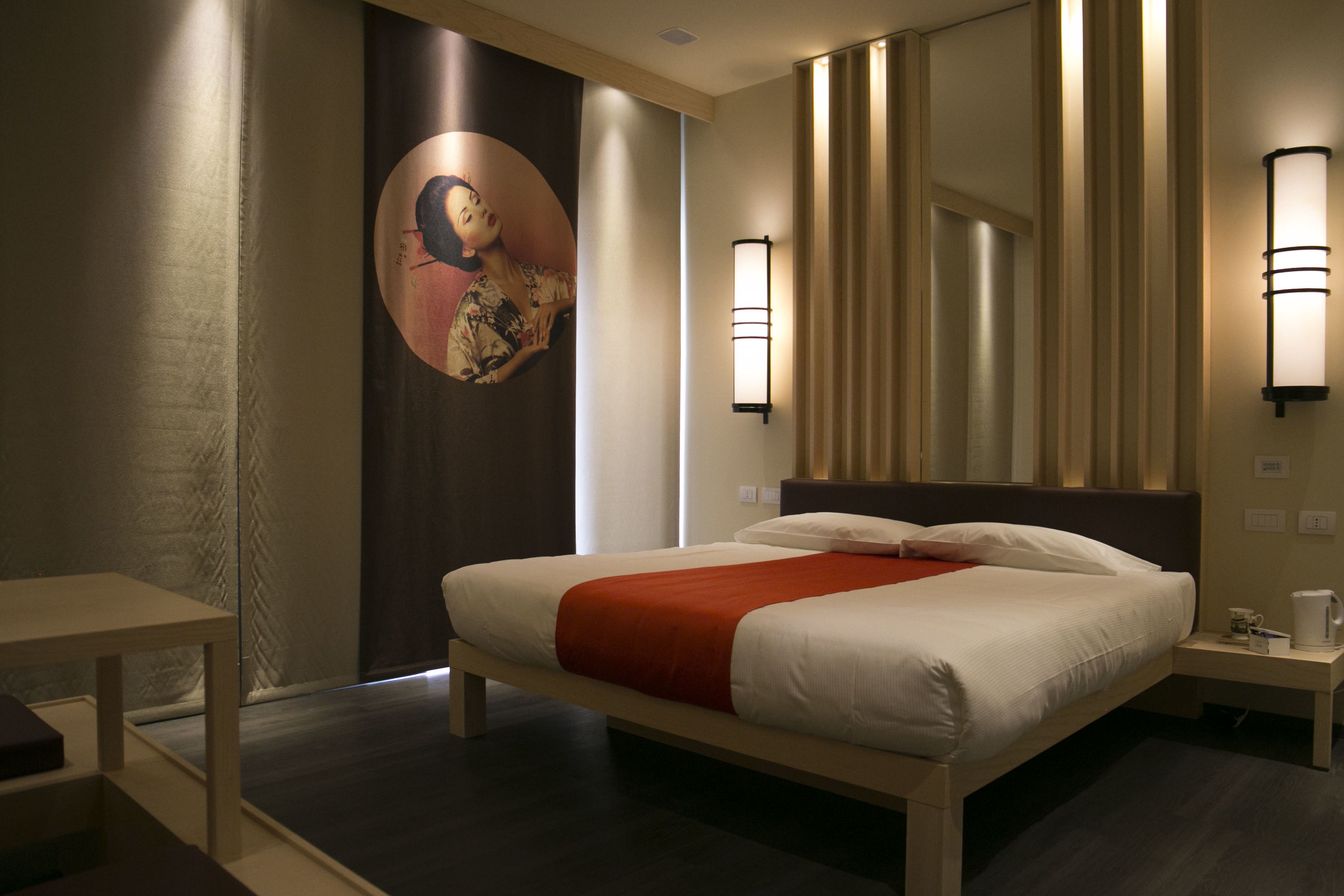 A hotel room with a bed, a nightstand, a wall-mounted lamp, and a large framed artwork.