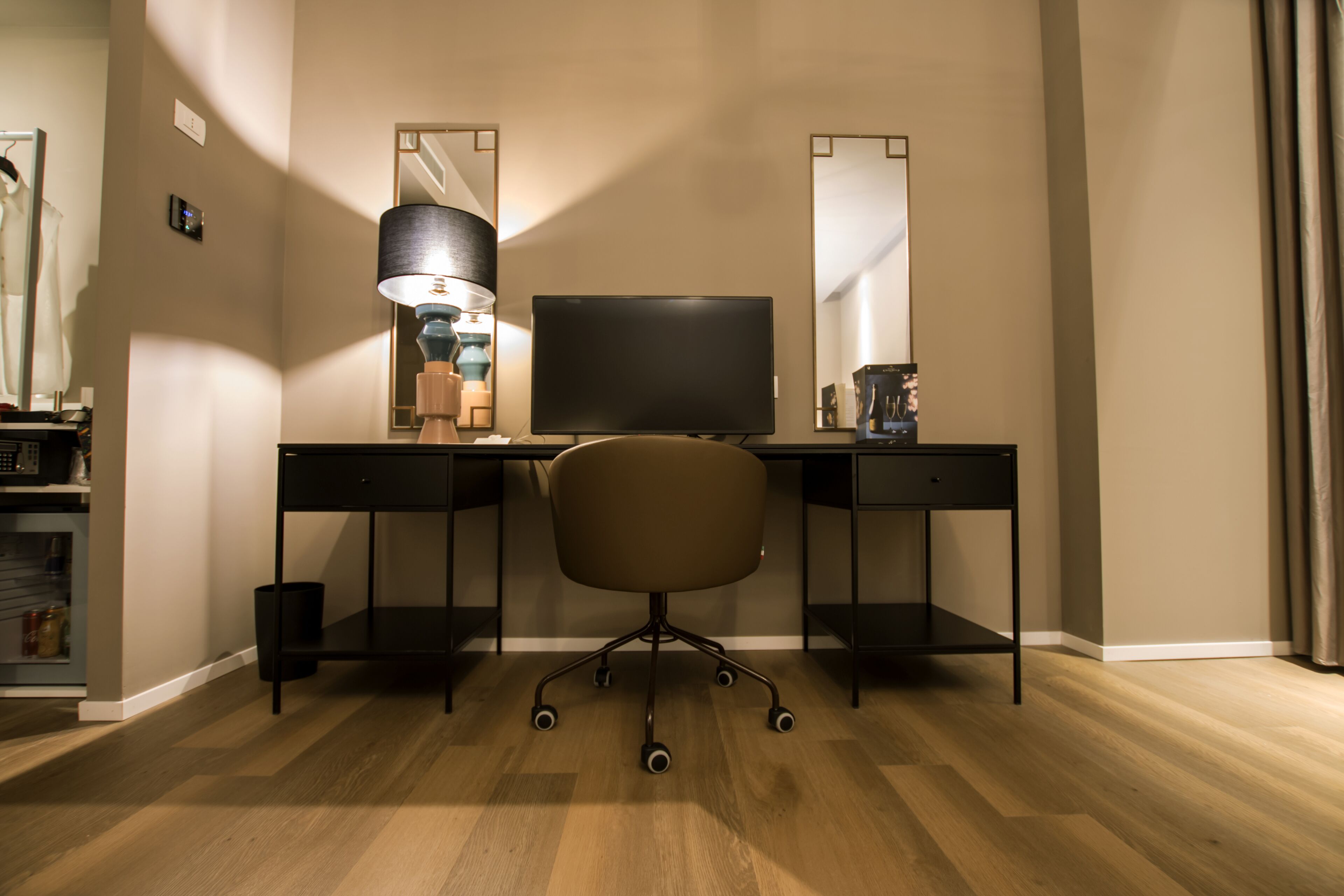 A modern hotel room with a wooden floor, a desk with a flat-screen TV, a chair, a lamp, and a mirror.