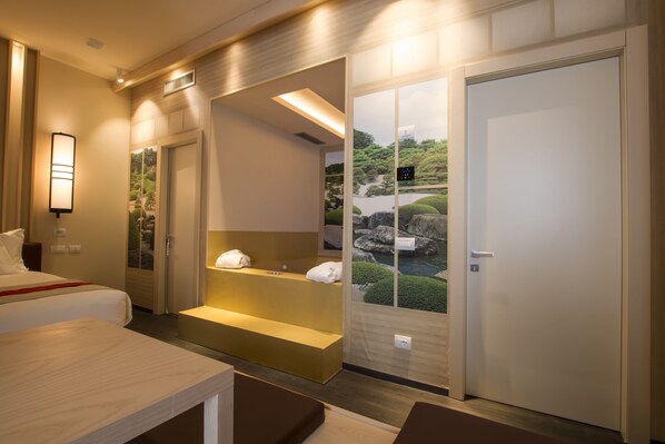 Habitación de hotel con cama, un pequeño rincón de descanso y vistas a un jardín, además de una puerta que da a otra habitación.